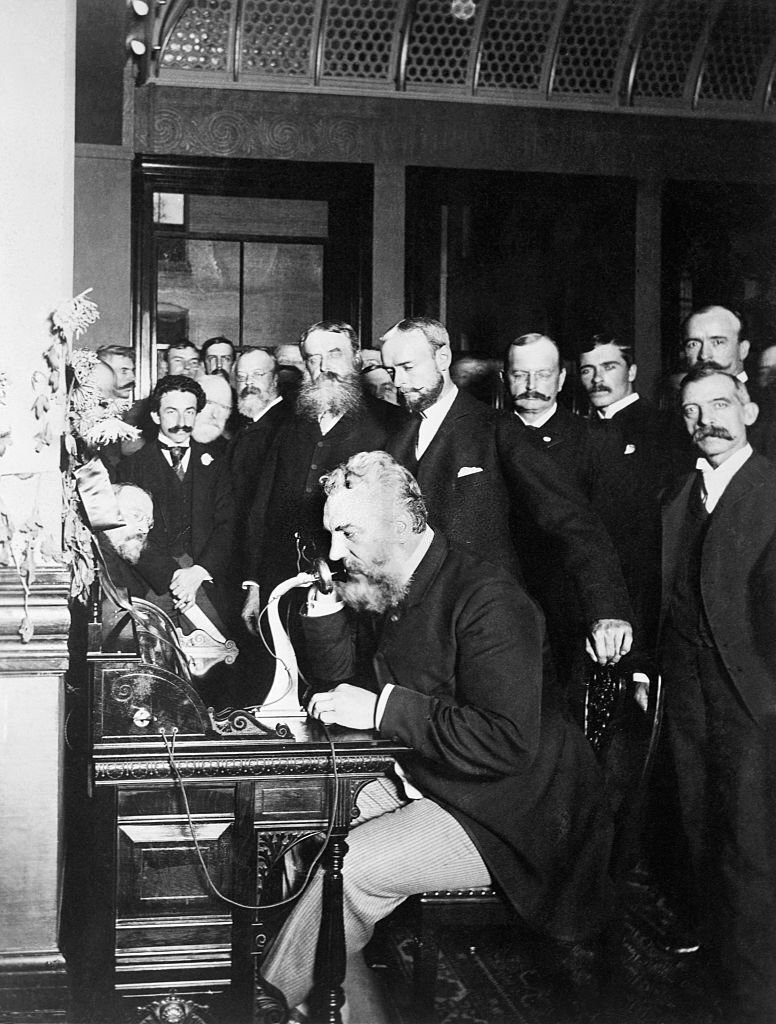 A group of businessmen watch inventor Alexander Graham Bell as he opens the New York-Chicago telephone line.